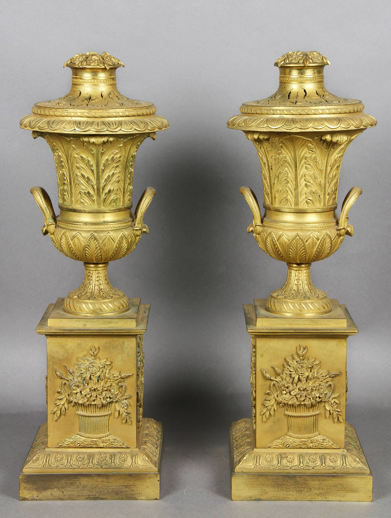 Each with a flower and leaf finial and pierced cover, the whole reversible to expose a candleholder over a campagna form two handled urn decorated with leaf tips and acanthus, the square form base with basket of flowers cast decoration.