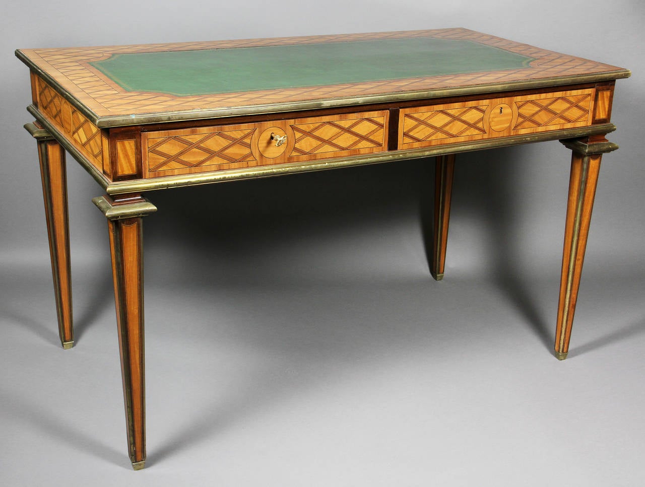 Rectangular top with trellis pattern inlay and inset tooled green leather top with brass edge over two drawers raised on square tapered legs with brass capitols and edging. In the French taste.