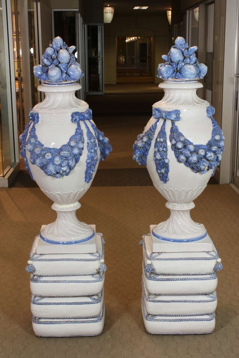This impressive pair of French blue and white glazed vases feature swags of fruit on the bodies, secured by bows, and separate lids ornamented with various fruits.  The vases rest on ceramic plinths in the form of pillows, which are most likely
