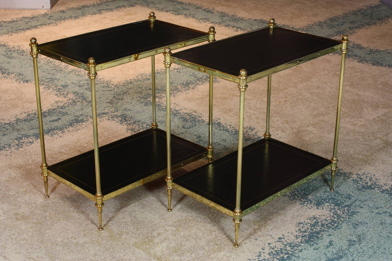 A handsome pair of French gilt brass two-tiered side tables with black leather tops, ball finials, fluted legs and tapered feet (attributed to Maison Jansen, mid-20th century).