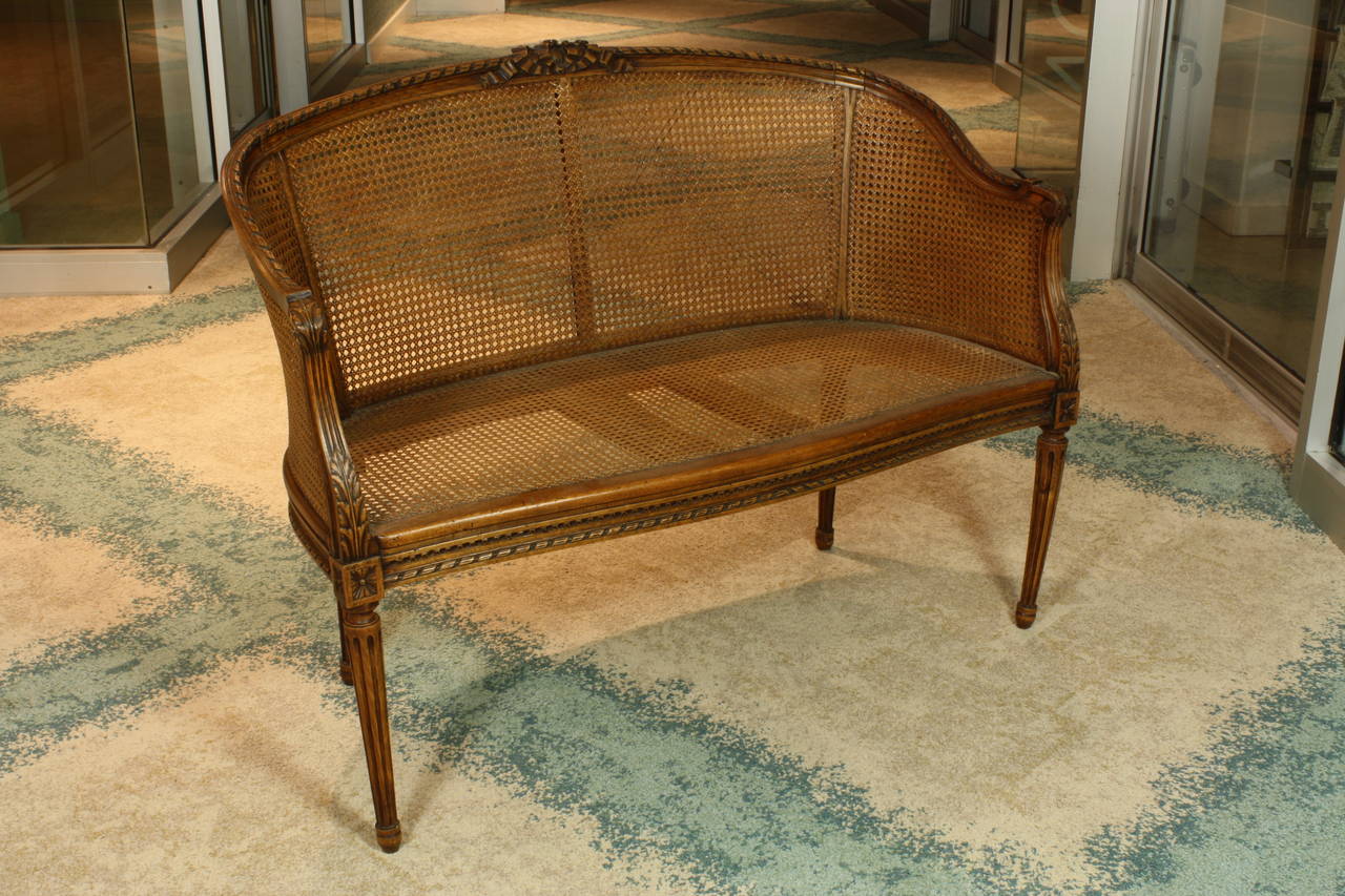 A pretty French Louis XVI style fruitwood settee or canapé with caned seat, back, and double-caned sides (Circa 1900).  The crest rail on the back has nicely-carved neoclassical bow, with carved ribbon and acanthus detailing along the frame and
