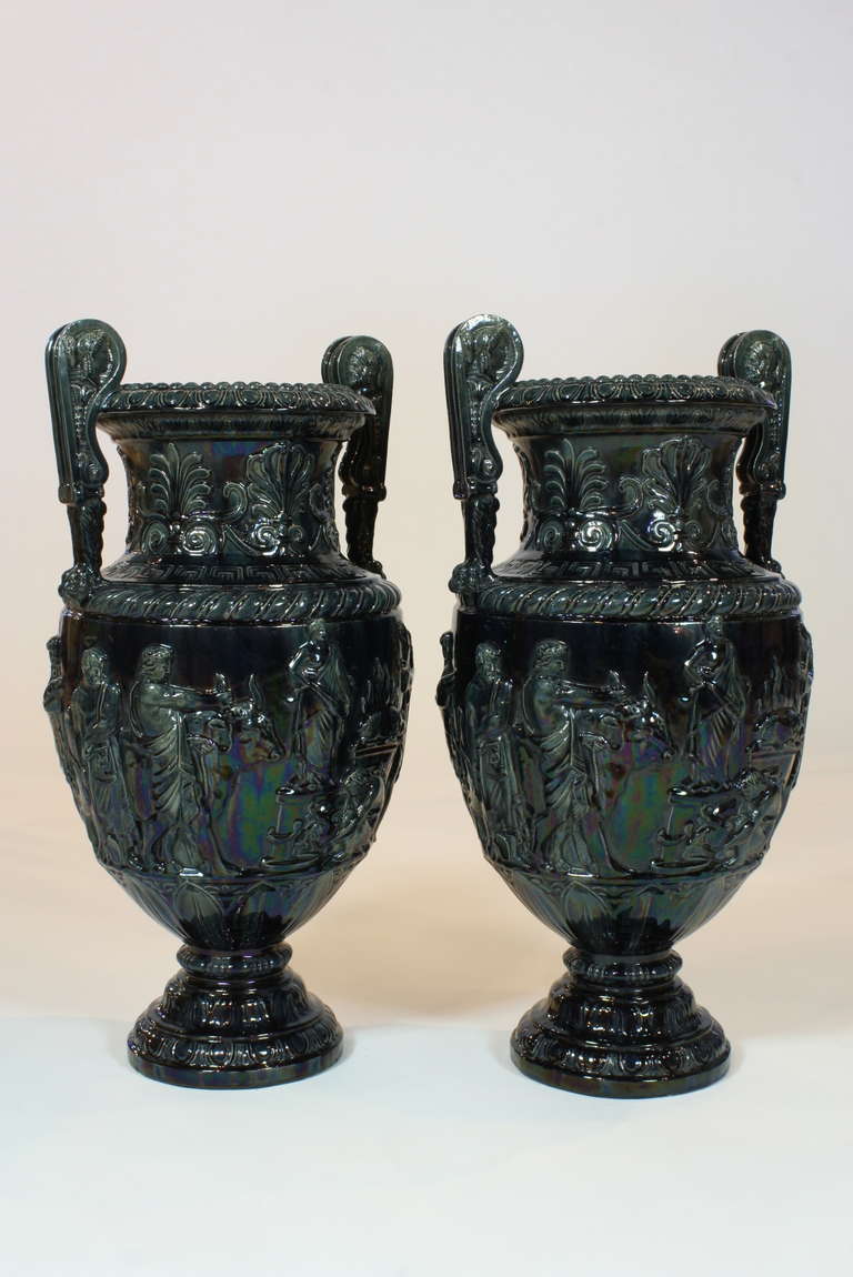 Impressive pair of late 19th century French neoclassical style ceramic urns with a deep blue-green iridescent glaze, in the form of a Grecian Volute Kraters.  The sides are decorated with scenes in relief of a festival, including figures playing