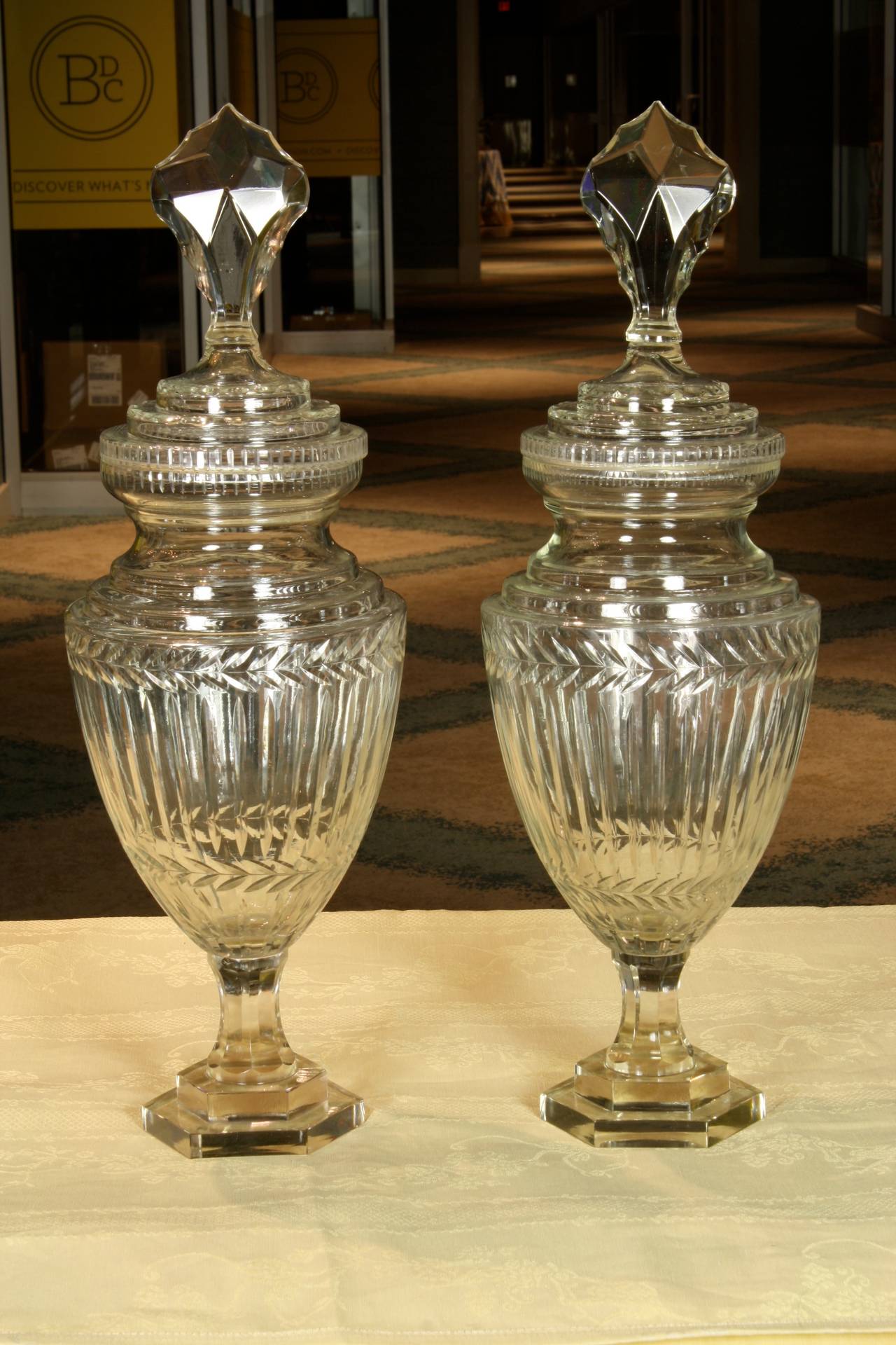 A pair of English hand-blown and cut-glass sweetmeat jars or urns, with glass covers topped with large cut-glass prism finials (Circa 1880).  Nice cut-glass leaf and other refined neoclassical decoration.
