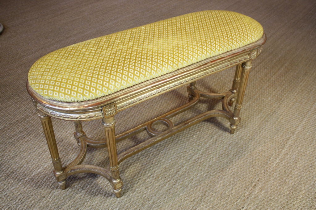 Elegant French giltwood bench in the Louis XVI style, with tapered fluted legs, carved ribbon detailing around the seat, pearl beading along the stretcher, and carved acanthus leaves, rosettes and other neoclassical elements. Newly-upholstered in