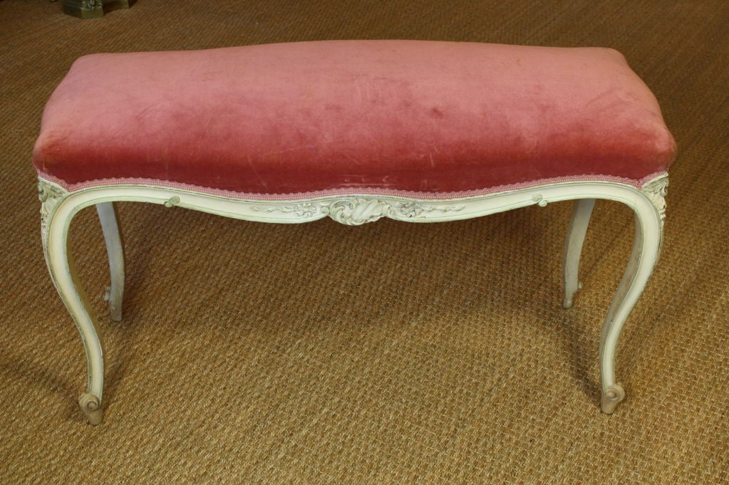French painted and upholstered bench in the Louis XV style, with cabriole legs, carved roses and floral detailing, some pale green highlights, and salmon-pink velvet fabric.