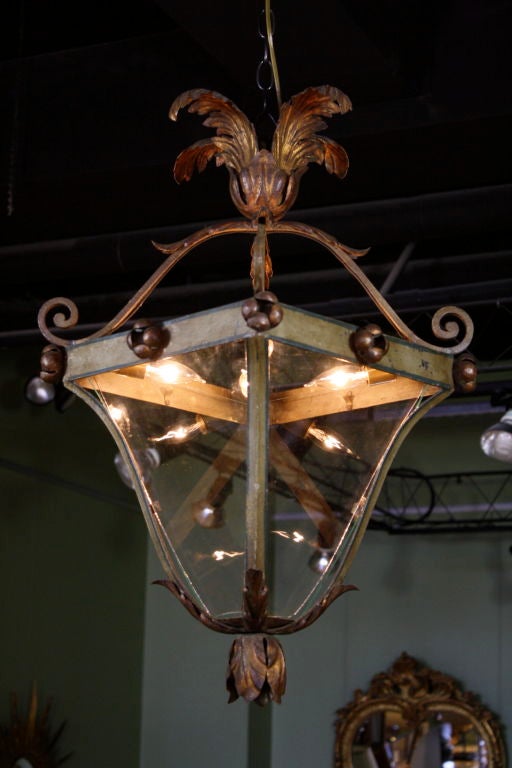 Italian iron lantern painted a dark green with gilded floral tole leaf elements. Scrolling wrought iron support on top, surmounted by gilded acanthus leaves; blossom shapes decorate the upper rim; and gilded acanthus leaves hang from the bottom of