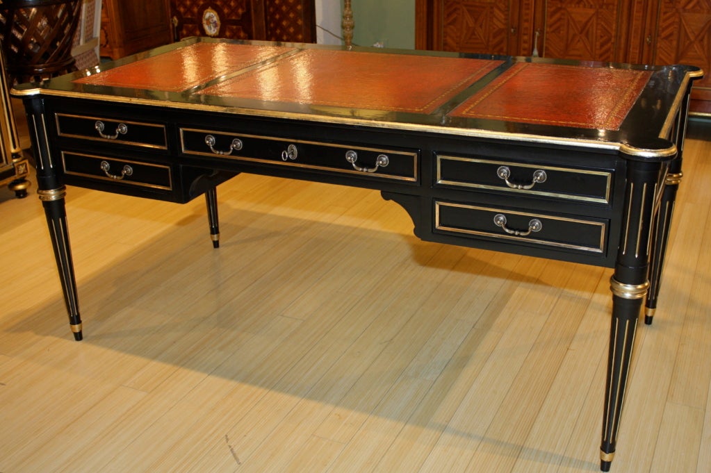 Louis XVI style black-lacquered desk with brass mounts and inlay, fluted legs with parcel gilt, embossed red leather top.  The desk is double-faced and features five drawers (the main central drawer has a working lock and key), and pull-out shelves