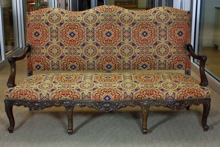 A nicely-carved French walnut settee or canapé with serpentine back, nicely-sculpted arms, seven legs (Regence style, circa 1870), upholstered in a Baroque style fabric with nail head trim. Carving features stylized scallop shells, acanthus leaves,