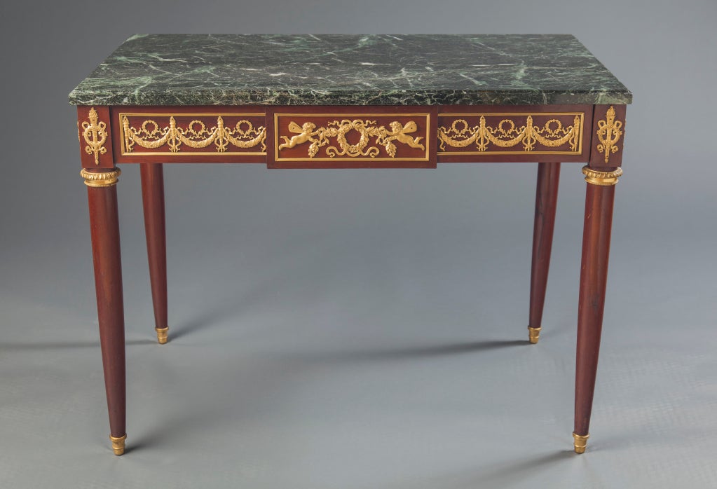 A French mahogany desk in neoclassical style with green variegated marble top, finely-chased gilt bronze mounts, featuring garlands of roses, torcheres and wreathes and putti with butterfly wings. Desk has one drawer. Stamped 