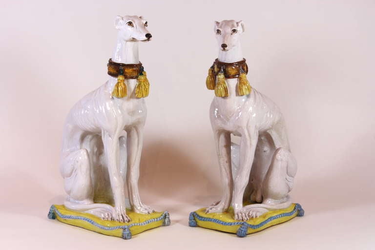 A pair of large Italian glazed ceramic grey-hounds with lovely buckled and tasseled collars, representing a male and female, seated on tasseled cushions.