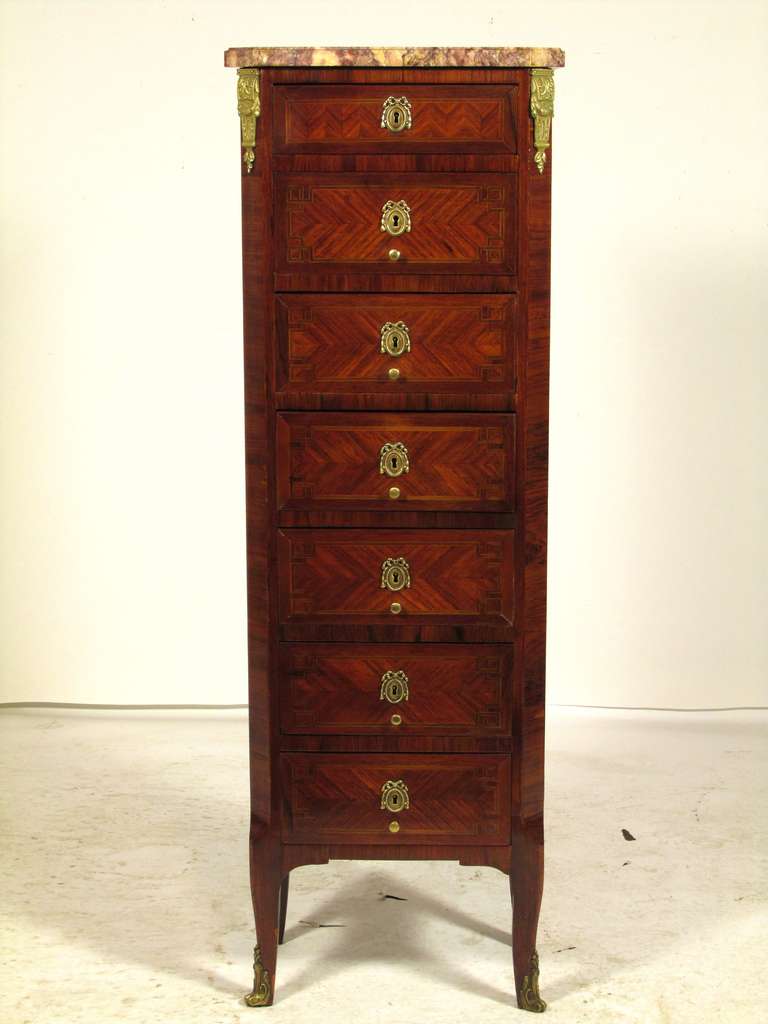 The French word for week is semaine.  A semainier is a tall narrow chest which must have seven drawers, one for each day of the week.  This semainier has veneers of kingwood and rosewood with a marble top.  The escutcheons and sabots are gilt bronze.