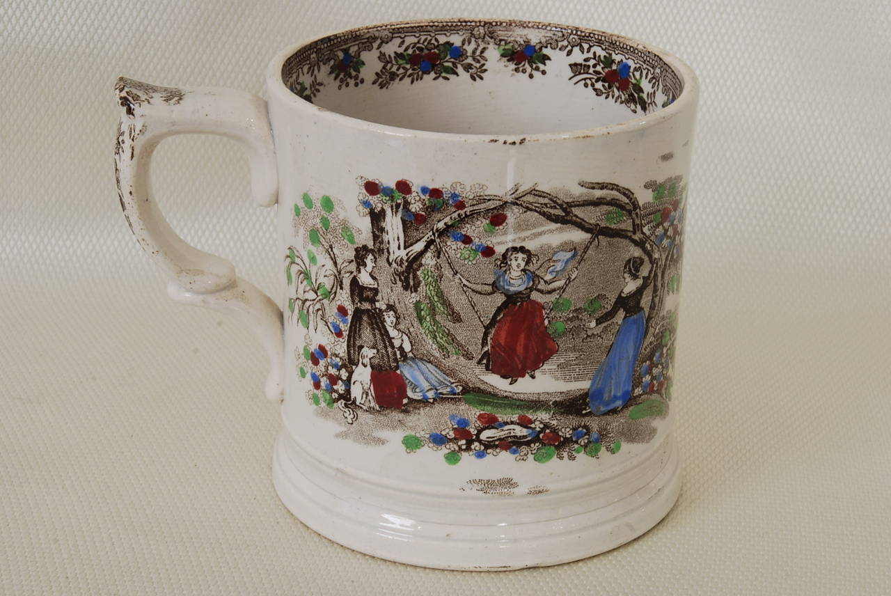 Staffordshire frog mug transfer printed in brown and over painted by hand. One side depicts fruit picking while the other side shows young ladies on a swing in a garden. The top inside rim has a transfer and hand painted border of flowers. The