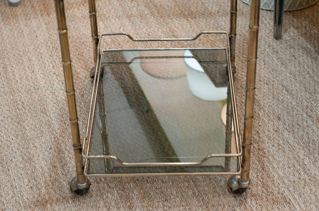 A vintage bar cart in brass and chrome with two tinted glass shelves on casters.