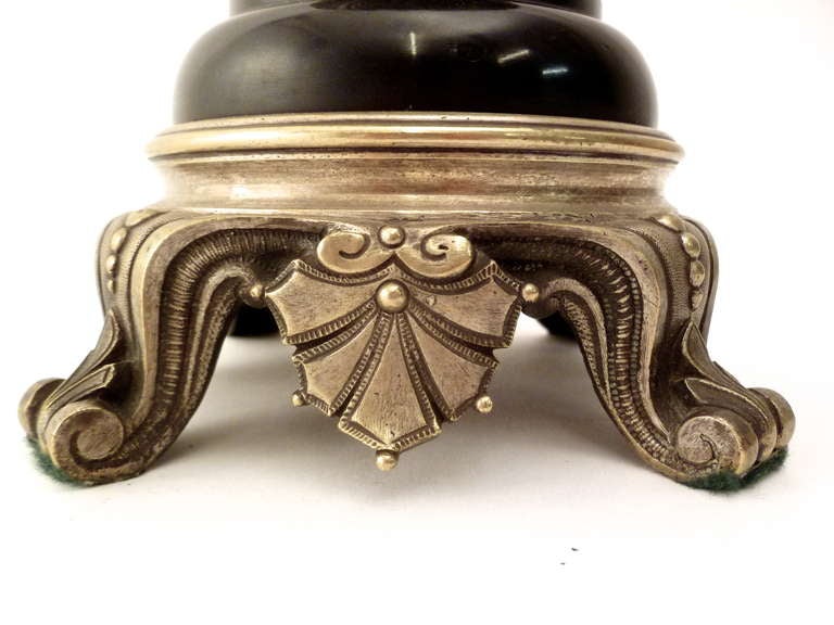 Delicate scrolling silvered bronze handles flank the slate body of the urn, with the sinuous aspect continued by an anthemion motif surrounding the waisted body. The base is supported by similarly scrolling feet and a repetition of the honeysuckle