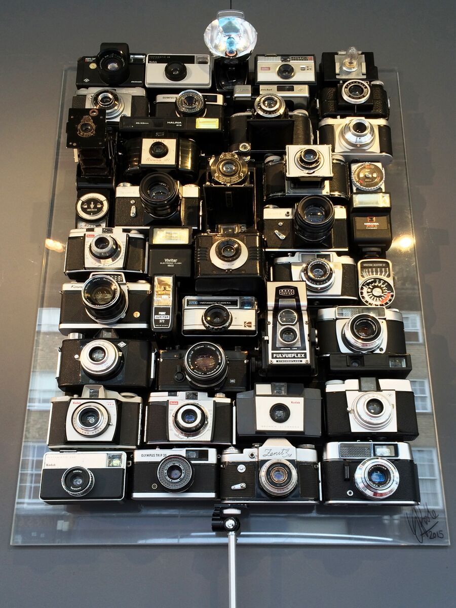 Eine Zusammenstellung von alten Foto- und Filmkameras, Blitzgeräten und Belichtungsmessern, montiert auf einer Platte. Das Kunstwerk ist an der Wand befestigt und in einer Plexiglasvitrine eingeschlossen. Ein altes Stativ ragt aus dem Boden der