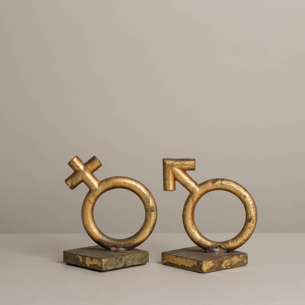 A pair of worn gold leafed cast metal bookends by Curtis Jere signed and dated '69 depicting the gender glyphs.

