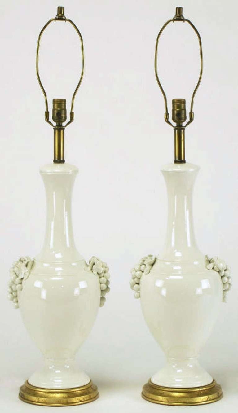 Elegant and early pair of urn form white ceramic table lamps with grape cluster handles from Frederick Cooper Studios. Gilt base with brass stem, socket and harp. Sold sans shades.