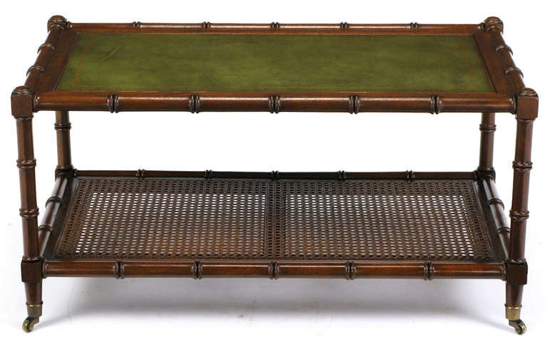 Mahogany, carved to resemble bamboo, framed rectangular petite coffee table. The top has a greened tooled leather insert and the bottom shelf is caned. The feet are finished in brass sabots and casters.