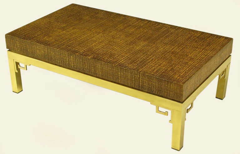 Table basse italienne avec plateau en bois recouvert de raphia et base en forme de clé grecque en laiton avec consoles. Le léger retrait de la menuiserie du plateau et de la base en laiton crée un effet de plateau flottant.