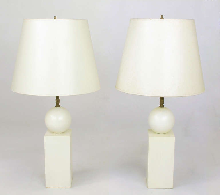 Uncommon pair of table lamps constructed of wood and in the  shape of a ball atop a pedestal. Off white lacquer is checking, adding more character. Double socket cluster and stem are made of brass. The shades are in fair condition, and we will