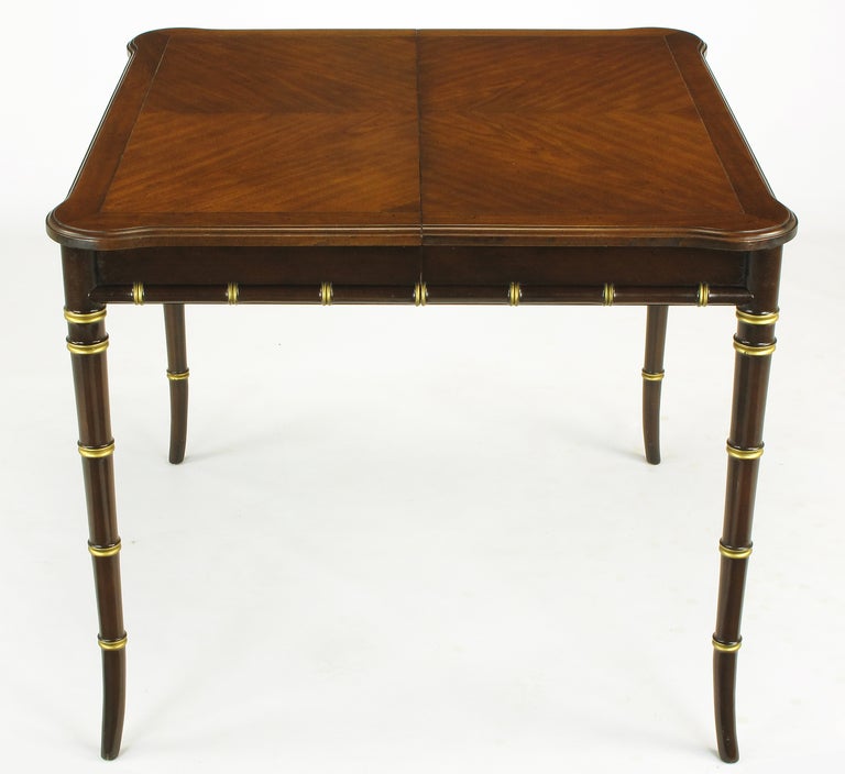 Excellent regency style petite dining table by Kindel Furniture. Deep oxblood lacquered base of stylized bamboo saber legs and apron edging with gilt banding. Book matched parquetry walnut top with rounded and extended corners and incised detail.