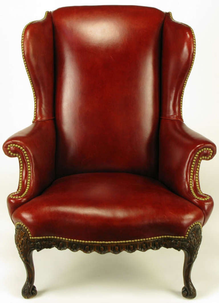 Red leather regency wingback chair with heavily carved walnut front legs and apron. Back legs are subtlely carved with a dramatic rake. Brass nail head detailing on the curvaceous arms, sides and apron.