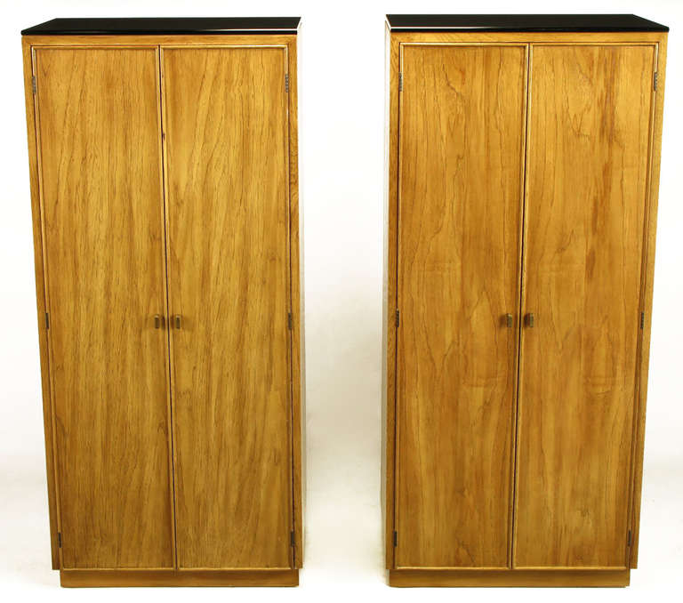 Pair of narrow tall cabinets in figured ash wood with factory applied patina. Black glass tops with bull nose edging and solid brass radius edge pulls. Two doors open to reveal four storage compartments. Would make a striking pair of tall night