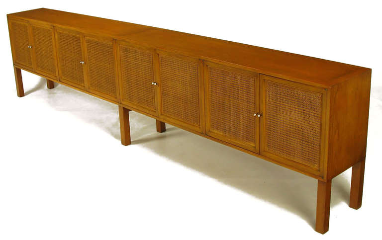 Exceptionally long and striking bleached walnut side board with woven cane door front panels and nickeled tear drop pulls. The eight doors open to reveal a single drawer section for flatware and shelved storage. Tall square dowel legs and the