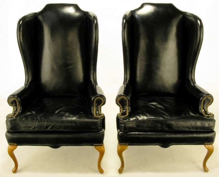 Pair of black leather upholstered wing chairs with a narrow visage and stately profile from Hickory Chair Company circa 1960s. Queen Anne style front legs and arched back legs are made of rock maple. Brass nail heads adorn the rolled arm fronts and