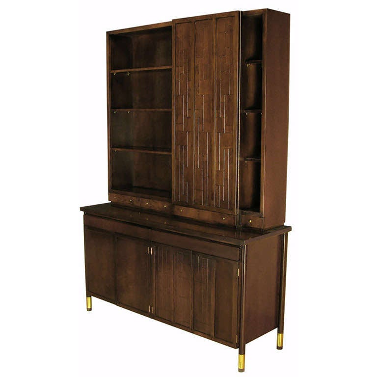 From Bert England's Forward Trend collection for Johnson Furniture, a two-piece tall cabinet in walnut with large dowel brass cuffed legs. Upper cabinet features three shelved sections, one section is behind a carved mosaic three panel door. Four