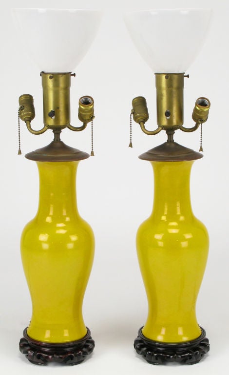Pair of Chinoiserie vase form table lamps with canary yellow glazed ceramic bodies. Bases are heavily carved wood bases with mahogany stain.  Triple socket illumination with center milk glass diffuser and pull chain switches. Sold sans shades.