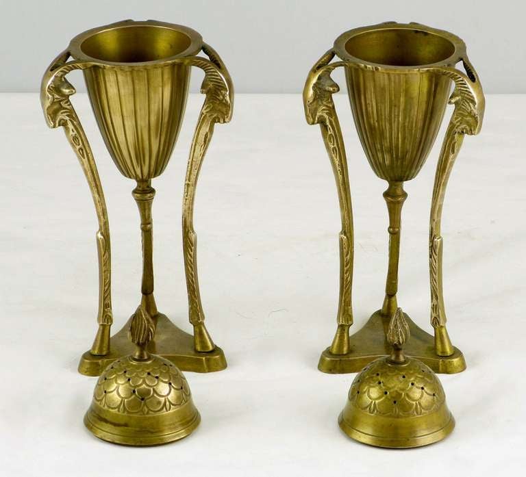 Beautifully patinated Empire styled brass urns adorned with three rams heads and hooved pilaster legs surmounted on trefoil brass bases. The pierced cap has a flame finial top and is removable. The conical incised center urn has a bottom finial. 