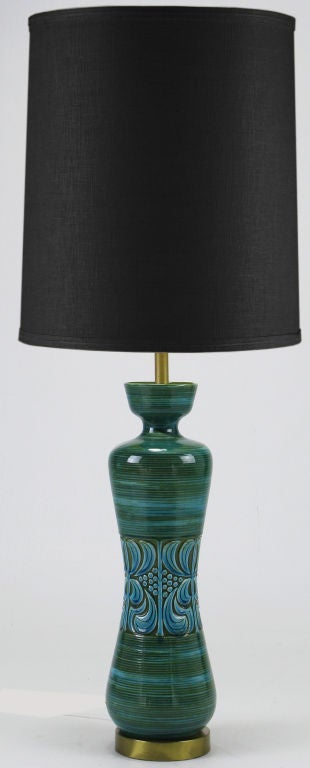 Hand thrown pottery table lamp in blue green glaze with brass base, stem, and socket. Hour glass shaped center, with impressed design, in the style of Bitossi for Raymor. Sold sans shade.