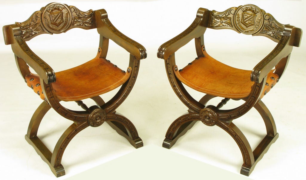 Pair of Gothic revival curule chairs in carved oak and umber leather with large brass nail head upholstery tacks. Hand carved backs feature square rosettes and center shields. X-shape sled bases feature center rosettes and carved stretchers.