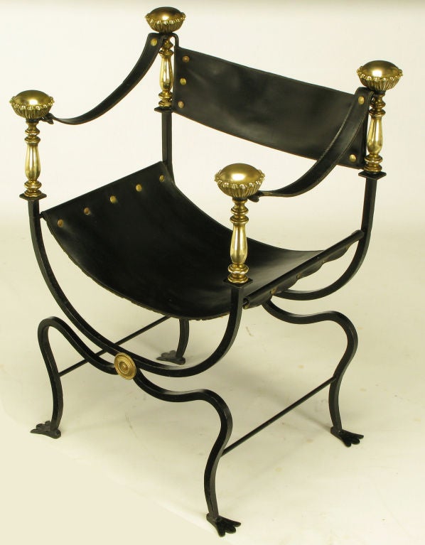 Black lacquered wrought iron stylized curule form chair with black leather sling seat, arms and back. Front and back U shaped supports feature oversized brass finials and brass spindles. Legs have stylized webbed feet.  Center brass rosette to the