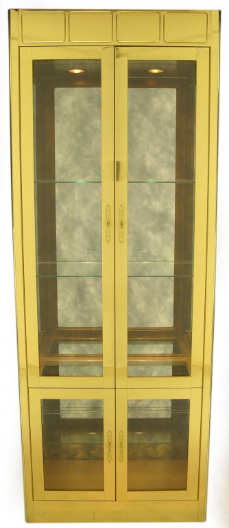 Mastercraft postmodern brass over wood four door vitrine with pierced escutcheons and open rectangular pulls. Taller upper section has two glass shelves and lower section has a single glass shelf. Aged silver mirror backing. Illuminated by two