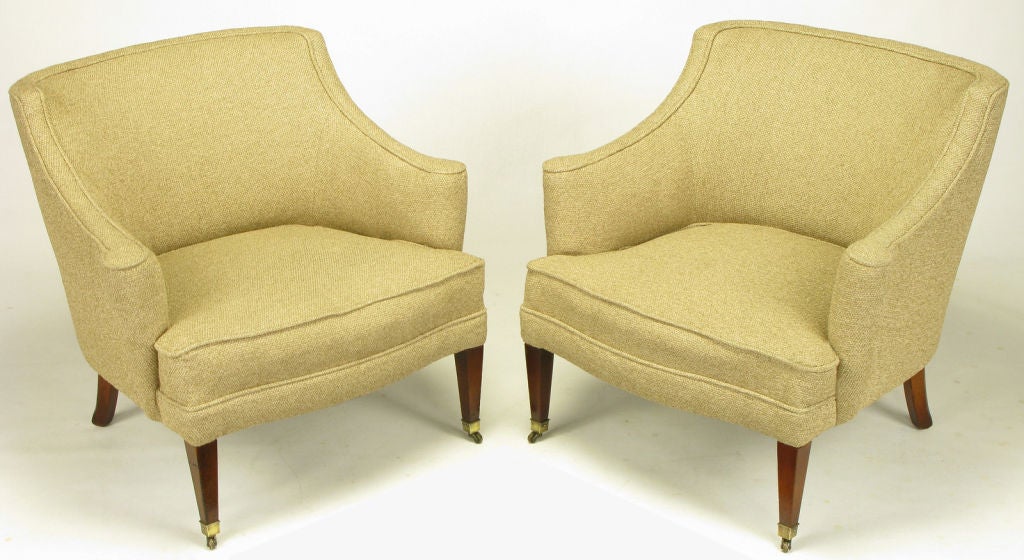 Pair of sleek 1940s rolled side arm club or lounge chairs with clean lined tapered mahogany legs and front casters. The upholstery is a vintage taupe woven wool.  Design & construction on par with Dunbar or Baker.