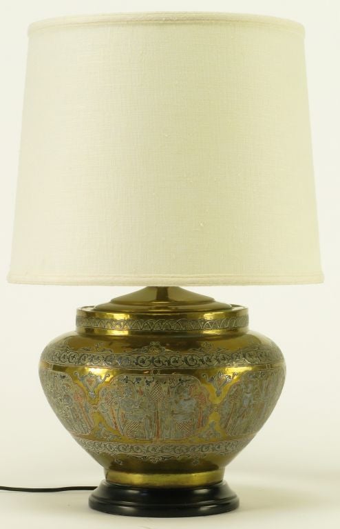 Urn shaped brass bodied table lamp with embossed and appliqued copper and pewter ornament. Egyptian scene with filigree borders and detailing. Acid etched brass reveals the copper under coat present on the figures and the filigree. Black lacquered