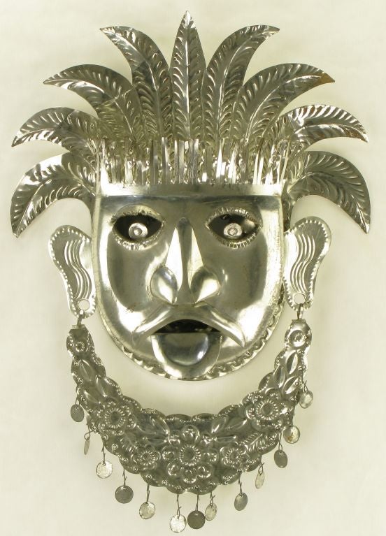 Stamped and cut polished metal tribal mask. Foliate headdress, pierced and plated lower lip with pierced ears joining together in a necklace. Three dimensional with lacquered black backed recessed eyes and mouth.