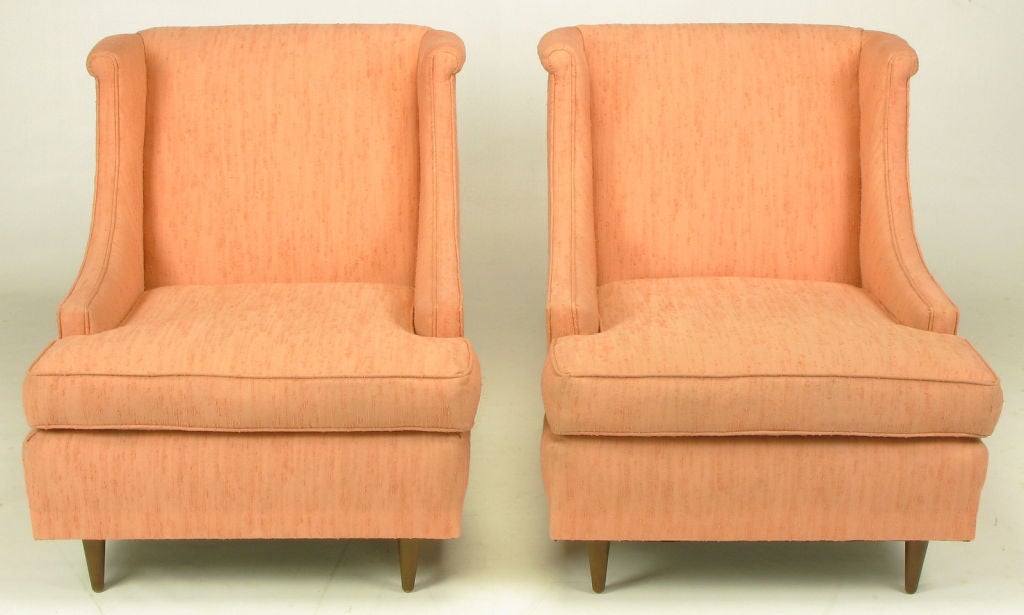 Pair of coral textured silk blend upholstered low wing chairs with deep seats and a rolled top back and wing. Low sloping arms give the feel and appearance of a slipper chair. Conical walnut legs.