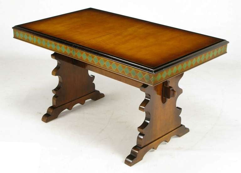 Maple trestle table with heavily carved legs and high, through-tenon trestle. Flame maple top has hand rubbed finish, gilt and black lacquered border, with a gilt and emerald green harlequin patterned apron. Internally stored single leaf.