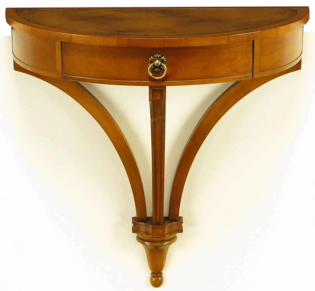Demilune light walnut and tooled leather wall mount console table. Three legs meet at a center carved sconce or bracket. Single center drawer with brass drop ring pull and escutcheon. Aged and tooled leather top.