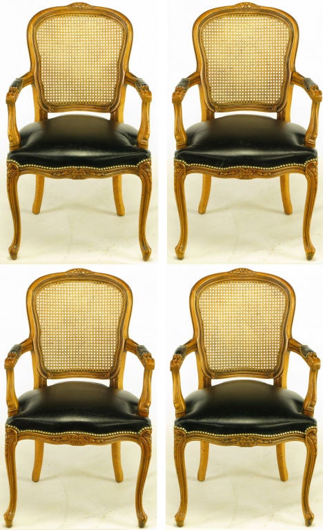 Set of four Louis XV style carved fruitwood arm chairs with black leather seats, arm pads and caned backs. Ornately carved apron, turned feet and arms. Finished in a light cherry antiqued glaze with speckled worm hole patina. Brass nail head