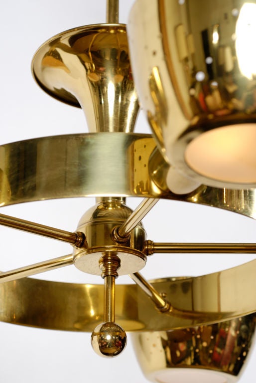 This stunning chandelier features an inverted brass trumpet atop the central body, which has five straight arms connected by a wide vertical band of brass. At the end of each arm is a spun brass shade with reticulated perforations and a frosted