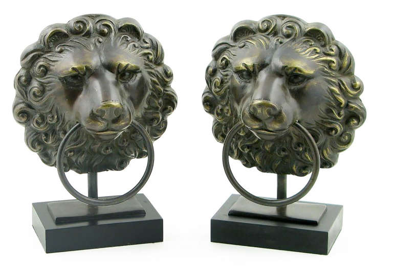 Backlit with candelabra bulbs, these two lamps feature cast lion heads mounted to a column upon a black plinth base. Each of the lions is clenching a large drop ring pull in its mouth.