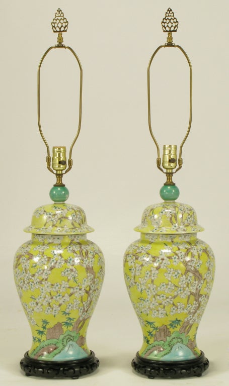 Pair of hand painted yellow glazed ceramic ginger jars with green finial table lamps on carved and ebonized wood bases. Hand painted white cherry blossoms on branches with a stream, castle, and foliate detail. Brass stem, harp, socket and finials.