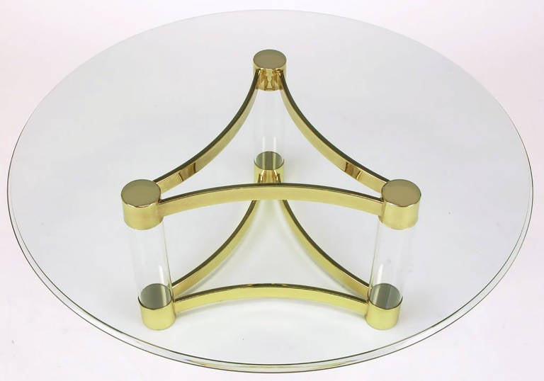 Three brass capped and footed Lucite tubes, connected by bent flat bar brass stretchers, combine to form a reverse trefoil coffee table base. Ogee edge round glass top is three-quarters of an inch thick.