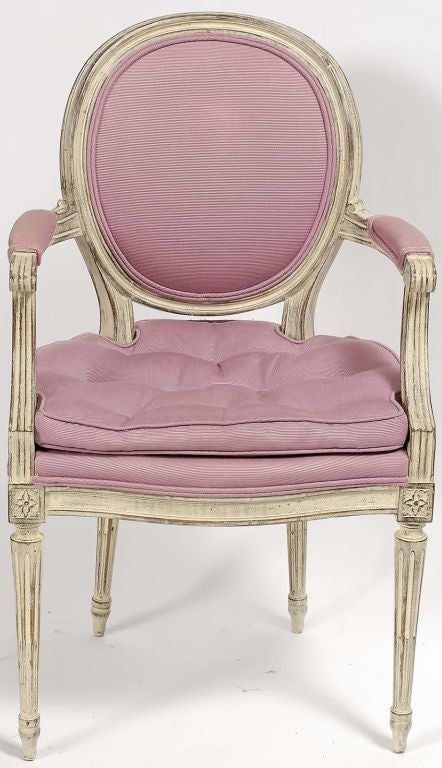 These chairs are a twentieth-century interpretation of a classic Louis XVI fauteuil. They are carved with rather distinctive flourishes. The finish is a rusticated ivory glaze over wood, and they are upholstered in a lavender twill fabric. 

The