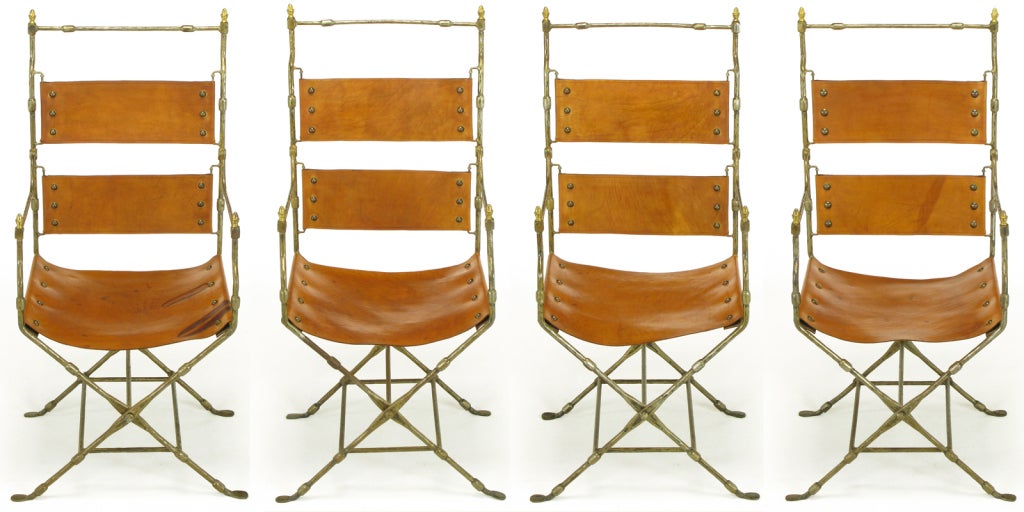 Set of four custom crafted hand forged iron and leather sling chairs with brass finials. Thick umber tanned saddle leather sling seats and two part back riveted in hand forged iron X-base frames with segmented construction and lower box stretcher.