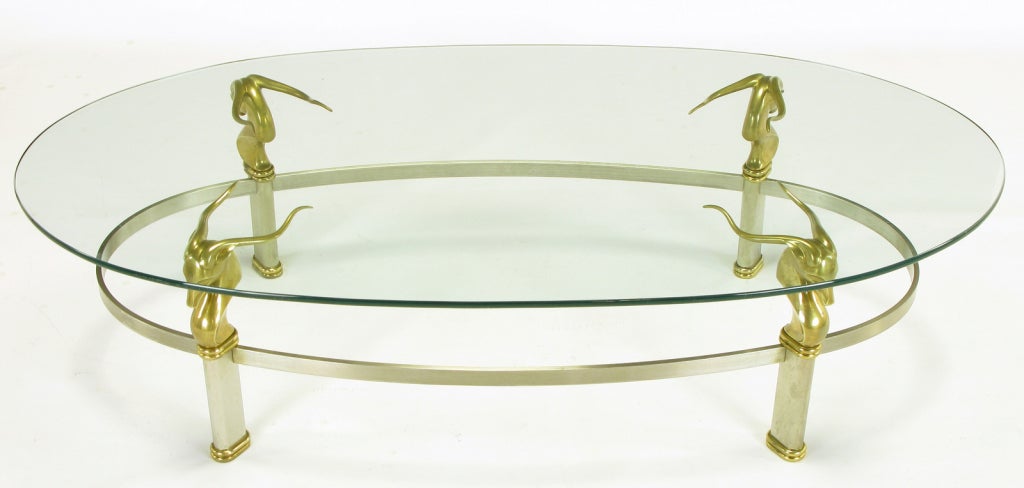 Table basse de forme ovale de style Art Déco, construite en acier inoxydable brossé, avec un châssis ovale et des pieds inclinés. Têtes de gazelles en laiton avec double perlage ovale en laiton aux pieds. Le plateau en verre a une épaisseur de 3/8